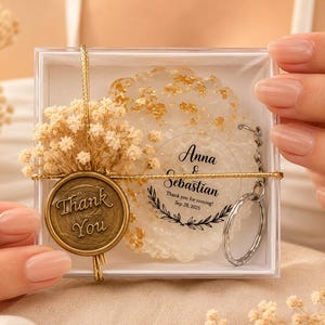 May include: A clear square gift box containing a round white resin keyring with gold flakes. The keyring features the text Anna & Sebastian, Thank you for coming! Sep 28, 2025. A gold wax seal with Thank You text and dried flowers are attached.