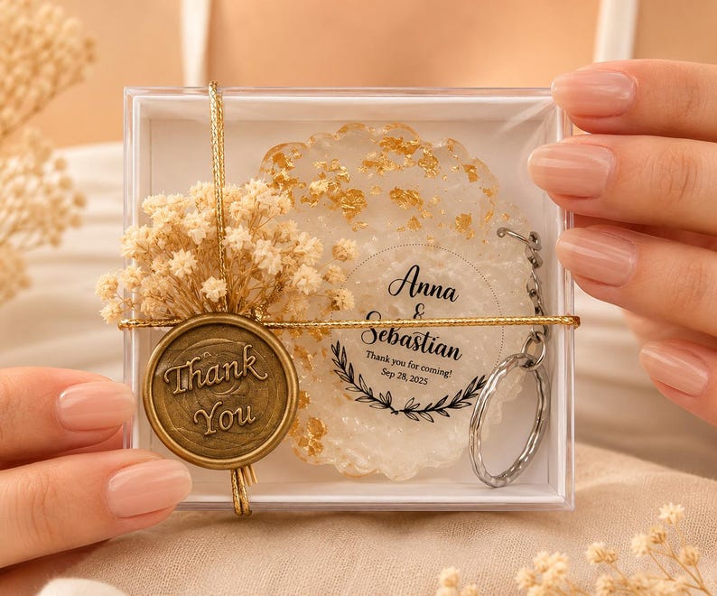 May include: A clear square gift box containing a round white resin keyring with gold flakes. The keyring features the text Anna & Sebastian, Thank you for coming! Sep 28, 2025. A gold wax seal with Thank You text and dried flowers are attached.