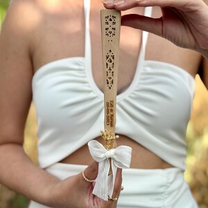 May include: A wooden fan with intricate cut-out designs. The fan has a white ribbon and dried flowers attached. The text "Ed & Bernat 09-09-2023" is engraved on the fan. The fan is held up against a white dress.