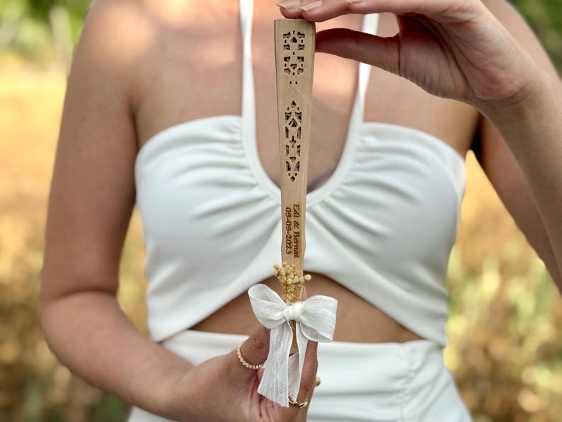 May include: A wooden fan with intricate cut-out designs. The fan has a white ribbon and dried flowers attached. The text "Ed & Bernat 09-09-2023" is engraved on the fan. The fan is held up against a white dress.
