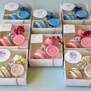 May include: A collection of square white gift boxes containing small round containers, dried flowers, and a wax seal stamped with the words Thank You. The boxes are tied with gold string and feature circular labels with various illustrations.