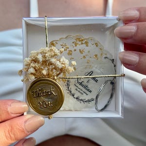 May include: A square white gift box containing a round resin coaster with gold flakes, a dried baby's breath flower bouquet, and a gold wax seal with the text Thank You. A circular tag reads Anna & Sebastian Thank you for coming Sep 28, 2025.