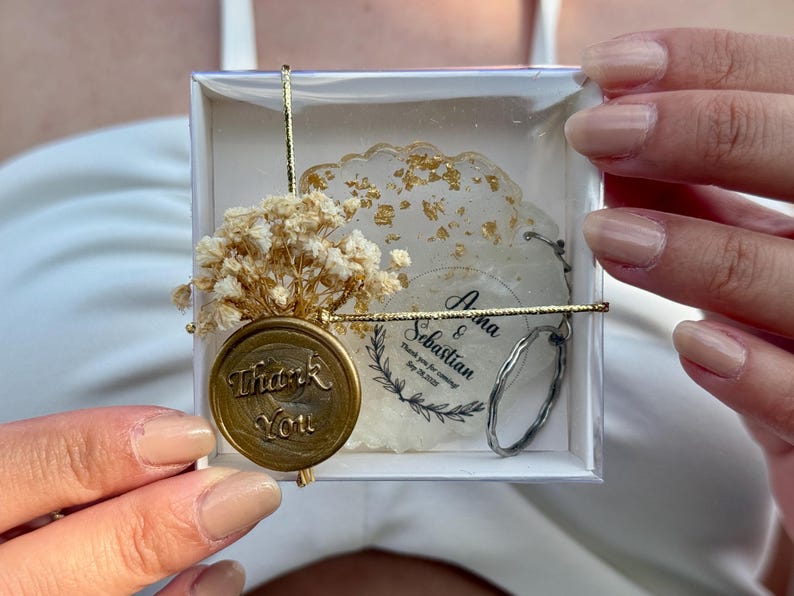 May include: A square white gift box containing a round resin coaster with gold flakes, a dried baby's breath flower bouquet, and a gold wax seal with the text Thank You. A circular tag reads Anna & Sebastian Thank you for coming Sep 28, 2025.