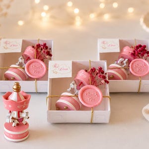 May include: Three white gift boxes containing a pink and white carousel ornament, a sprig of dried red flowers, and a pink wax seal with the text Thank You. A loose pink and white carousel ornament stands in front of the boxes with a gold top.