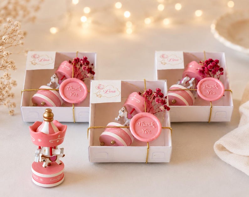 May include: Three white gift boxes containing a pink and white carousel ornament, a sprig of dried red flowers, and a pink wax seal with the text Thank You. A loose pink and white carousel ornament stands in front of the boxes with a gold top.