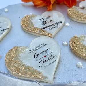 May include: Heart-shaped wedding favors with gold leaf accents and personalized text. Each favor features the names of the couple and the wedding date. The favors are arranged on a silver tray with pearl accents.