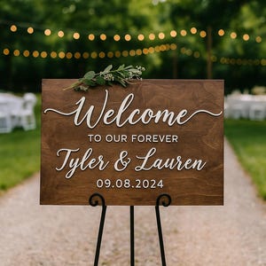 Puede incluir: Un letrero de bienvenida de madera con letras blancas que dice "Welcome to our forever Tyler & Lauren 09.08.2024". El letrero est&aacute; decorado con vegetaci&oacute;n y se muestra sobre un soporte de metal negro. El fondo presenta un entorno exterior borroso con luces de cadena.