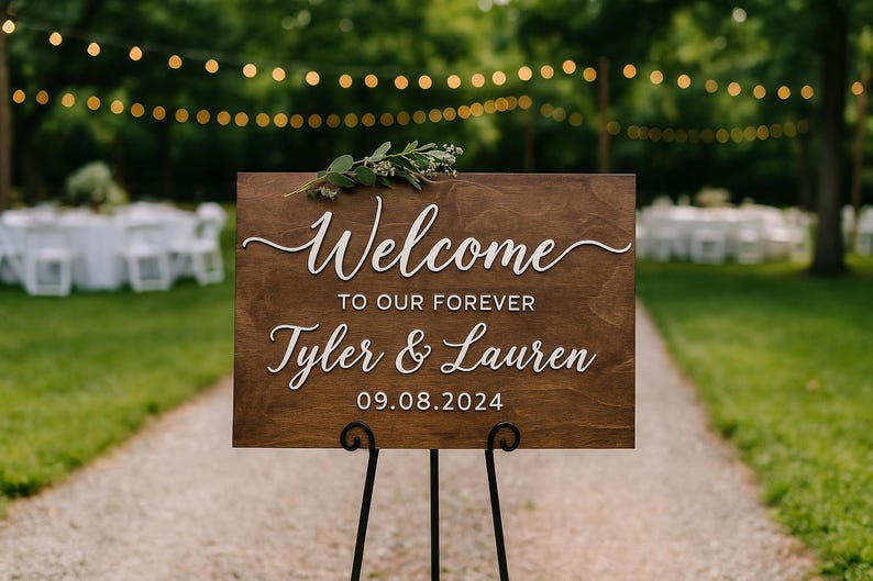 Puede incluir: Un letrero de bienvenida de madera con letras blancas que dice "Welcome to our forever Tyler & Lauren 09.08.2024". El letrero est&aacute; decorado con vegetaci&oacute;n y se muestra sobre un soporte de metal negro. El fondo presenta un entorno exterior borroso con luces de cadena.