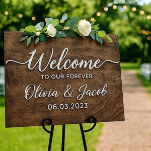 Puede incluir: Un letrero de bienvenida de boda de madera con las palabras "Welcome to our forever Olivia & Jacob 06.03.2023" en escritura blanca. El letrero est&aacute; decorado con rosas blancas y vegetaci&oacute;n y se muestra en un soporte de metal negro.