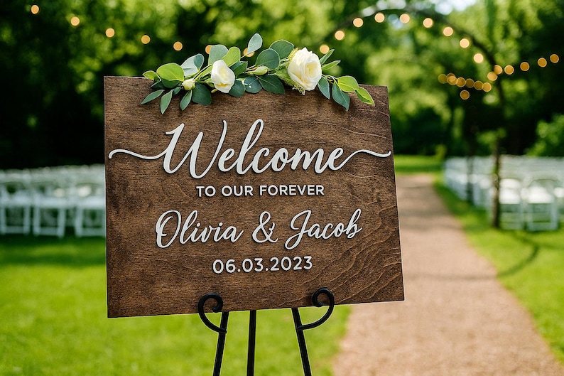 Puede incluir: Un letrero de bienvenida de boda de madera con las palabras "Welcome to our forever Olivia & Jacob 06.03.2023" en escritura blanca. El letrero est&aacute; decorado con rosas blancas y vegetaci&oacute;n y se muestra en un soporte de metal negro.