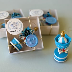 May include: Three white square gift boxes containing miniature blue and white carousel ornaments, each tied with gold string. A round sticker on the box features a cartoon elephant and the text Hoşgeldin Bebeğim, with a blue wax seal reading Thank You.