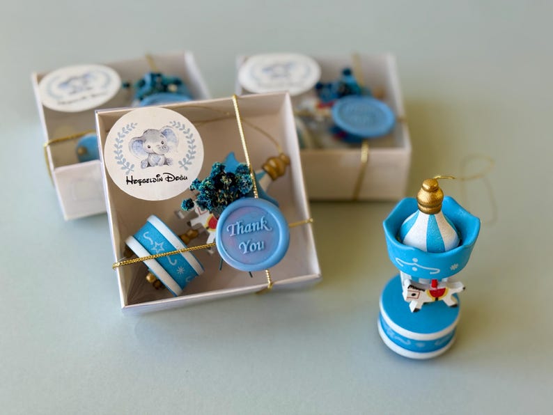 May include: Three white square gift boxes containing miniature blue and white carousel ornaments, each tied with gold string. A round sticker on the box features a cartoon elephant and the text Hoşgeldin Bebeğim, with a blue wax seal reading Thank You.