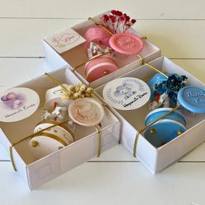 May include: Three white gift boxes containing small carousel-style music boxes in pink, white, and blue. Each box features a round sticker with illustrations of animals and balloons, a wax seal stamped with "Thank You," and dried floral accents.