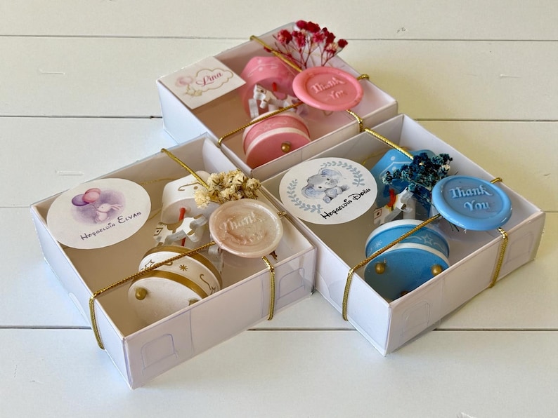 May include: Three white gift boxes containing small carousel-style music boxes in pink, white, and blue. Each box features a round sticker with illustrations of animals and balloons, a wax seal stamped with "Thank You," and dried floral accents.