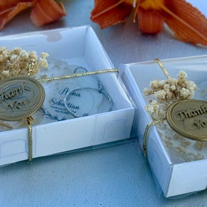 May include: Two square white gift boxes containing circular ornaments with the text "Nina Sebastian" and "Thank You" on gold wax seals. Each box is tied with gold string and decorated with a small cluster of dried white flowers on top.