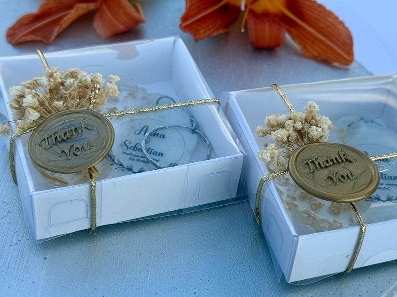 May include: Two square white gift boxes containing circular ornaments with the text "Nina Sebastian" and "Thank You" on gold wax seals. Each box is tied with gold string and decorated with a small cluster of dried white flowers on top.