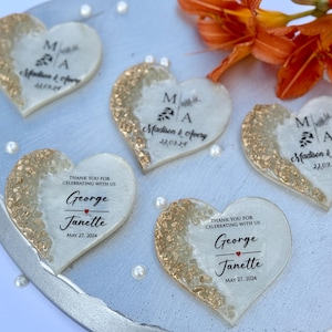 May include: Heart-shaped wedding favors with gold glitter accents and personalized text. The favors feature the names "Madison & Avery" and "George & Janette" with their wedding dates. Orange flowers and pearls are in the background.