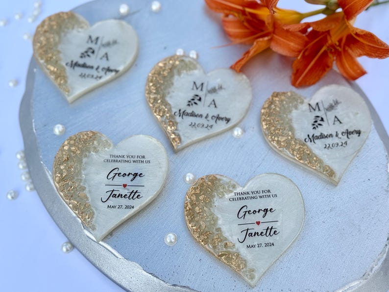 May include: Heart-shaped wedding favors with gold glitter accents and personalized text. The favors feature the names "Madison & Avery" and "George & Janette" with their wedding dates. Orange flowers and pearls are in the background.