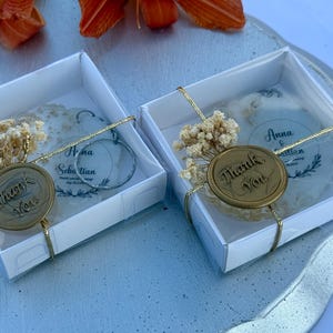 May include: Two square white gift boxes containing round translucent ornaments with the text Anna & Sebastian. Each box is tied with gold string, a sprig of dried baby's breath, and a circular wax seal embossed with the words Thank You.