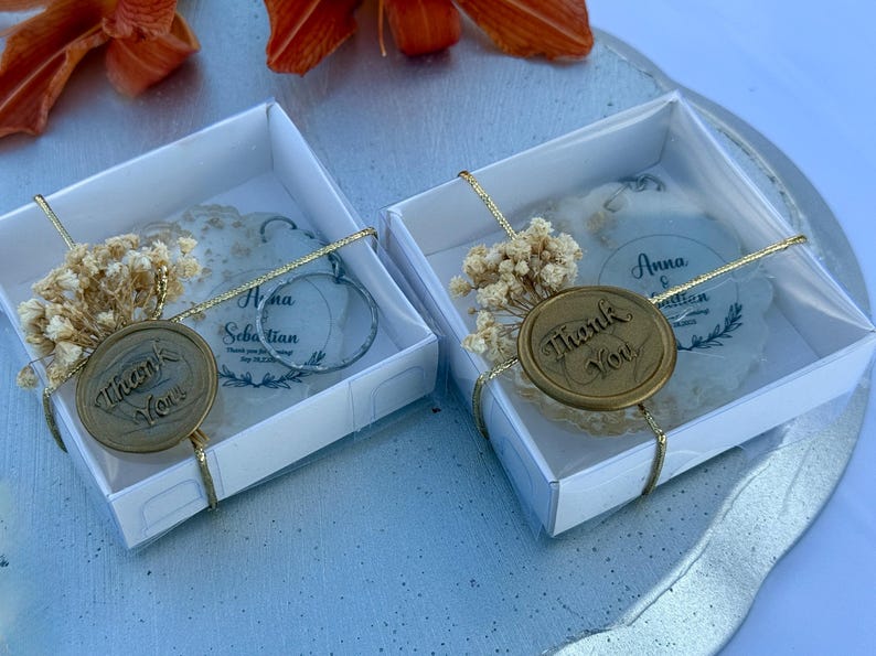May include: Two square white gift boxes containing round translucent ornaments with the text Anna & Sebastian. Each box is tied with gold string, a sprig of dried baby's breath, and a circular wax seal embossed with the words Thank You.