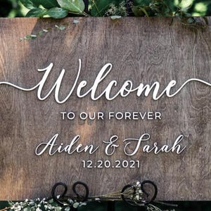Puede incluir: Cartel de bienvenida de boda de madera con las palabras "Welcome to our Forever" en escritura blanca. Tambi&eacute;n se muestran los nombres "Aiden & Sarah" y la fecha "12.20.2021". El cartel est&aacute; decorado con vegetaci&oacute;n.