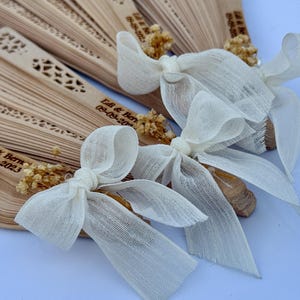 May include: Wooden hand fans with decorative cutouts and personalized text, tied with ivory fabric bows and dried floral accents. The fans are arranged in a fanned-out display, perfect for a wedding or special event.