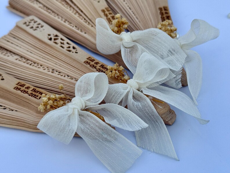 May include: Wooden hand fans with decorative cutouts and personalized text, tied with ivory fabric bows and dried floral accents. The fans are arranged in a fanned-out display, perfect for a wedding or special event.