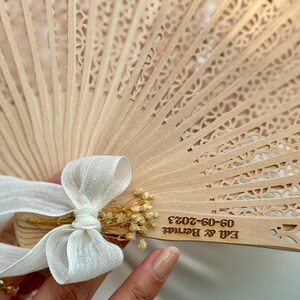 May include: A light brown wooden hand fan with intricate cut-out designs. The fan is adorned with a white ribbon and dried flowers. The text "Edi & Bernat 09-09-2023" is engraved on the fan, suggesting a wedding or special event.