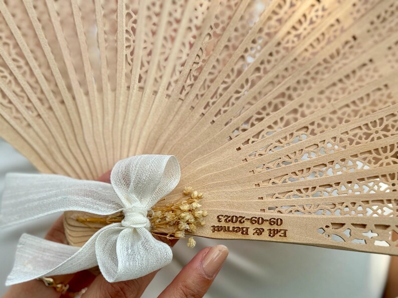 May include: A light brown wooden hand fan with intricate cut-out designs. The fan is adorned with a white ribbon and dried flowers. The text "Edi & Bernat 09-09-2023" is engraved on the fan, suggesting a wedding or special event.