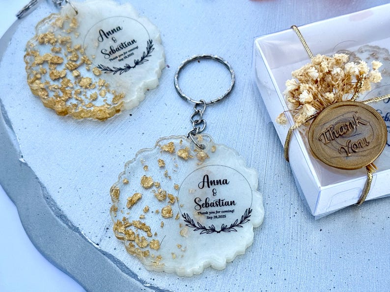 May include: Two round resin keychains with gold leaf flakes and white pigment. Each features black text reading Anna & Sebastian, Thank you for coming, Sep 28, 2025, framed by a laurel wreath. A white gift box with dried flowers and a Thank You wax seal.