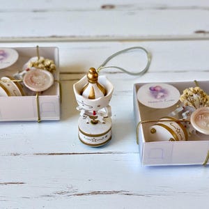May include: A white and gold carousel ornament with a hanging loop, positioned between two square white gift boxes. Each box contains a small carousel piece, a sprig of dried flowers, a round sticker, and a wax seal with the text Thank You.
