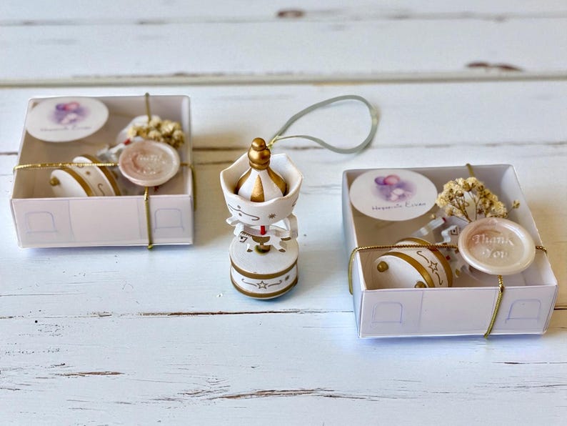May include: A white and gold carousel ornament with a hanging loop, positioned between two square white gift boxes. Each box contains a small carousel piece, a sprig of dried flowers, a round sticker, and a wax seal with the text Thank You.