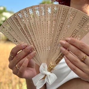 May include: A light brown wooden hand fan with intricate cut-out designs. The fan is held open, revealing its delicate pattern. A white ribbon and dried flowers adorn the base. Text is visible on the fan. Ideal for weddings or special events.