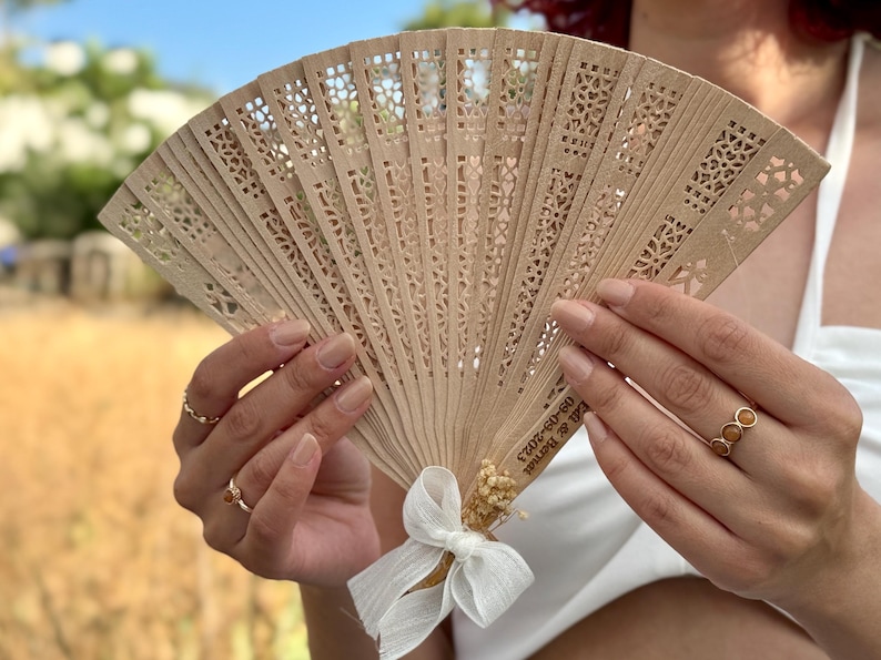 May include: A light brown wooden hand fan with intricate cut-out designs. The fan is held open, revealing its delicate pattern. A white ribbon and dried flowers adorn the base. Text is visible on the fan. Ideal for weddings or special events.