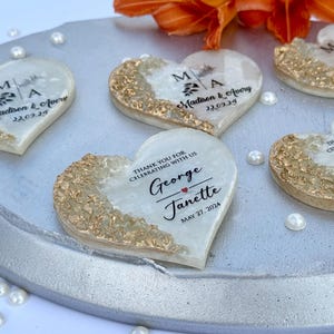 May include: Heart-shaped wedding favors with gold accents and personalized text. Each favor has a gold-colored edge and a white center with names and dates. The favors are arranged on a silver tray with scattered pearls and an orange flower.
