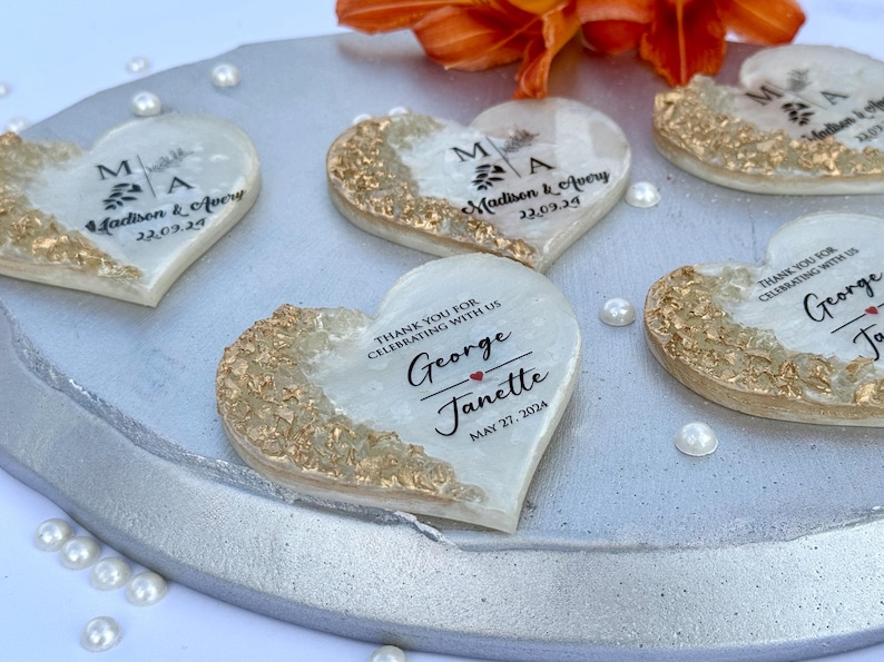 May include: Heart-shaped wedding favors with gold accents and personalized text. Each favor has a gold-colored edge and a white center with names and dates. The favors are arranged on a silver tray with scattered pearls and an orange flower.