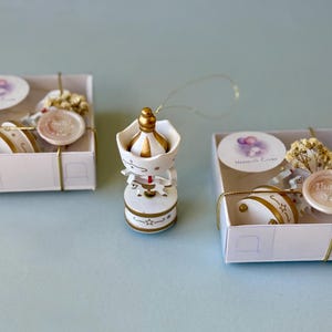 May include: A white and gold carousel ornament with a hanging string, positioned between two square white gift boxes. Each box contains a carousel ornament, a small sprig of dried flowers, a round tag, and a wax seal reading Thank You.