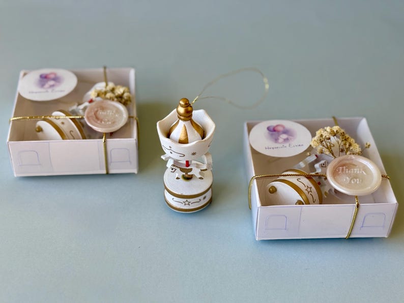 May include: A white and gold carousel ornament with a hanging string, positioned between two square white gift boxes. Each box contains a carousel ornament, a small sprig of dried flowers, a round tag, and a wax seal reading Thank You.