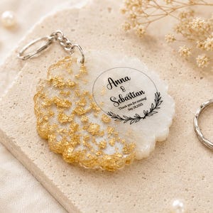 May include: A round white resin keyring featuring gold leaf flakes on one side and black printed text that reads Anna & Sebastian, Thank you for coming! Sep 28, 2025, framed by a circular border with a leaf motif and a silver metal ring.