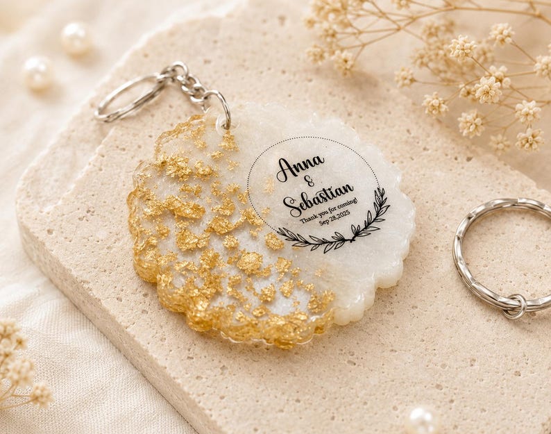 May include: A round white resin keyring featuring gold leaf flakes on one side and black printed text that reads Anna & Sebastian, Thank you for coming! Sep 28, 2025, framed by a circular border with a leaf motif and a silver metal ring.