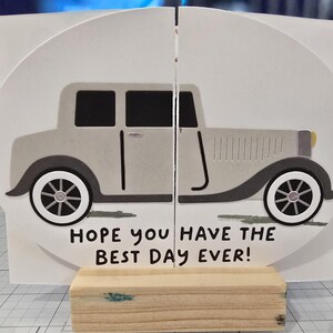 Può includere: Biglietto di auguri con un'illustrazione di un'auto d'epoca in tonalità grigie e bianche. Il biglietto, dalla forma unica, riporta la scritta "HOPE YOU HAVE THE BEST DAY EVER!". È sostenuto da un piccolo blocco di legno.