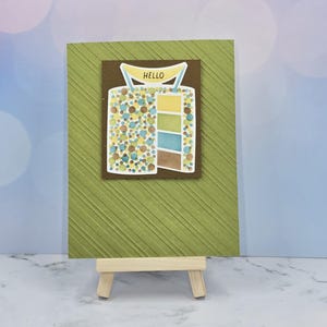 May include: A greeting card with a textured olive green background featuring a brown square overlay. The overlay displays a graphic of a cake decorated with colorful confetti dots and a yellow banner at the top with the word HELLO printed in black.