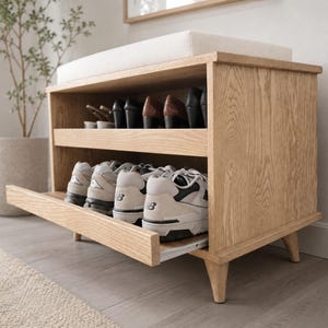 Può includere: Una panca scarpiera in legno di rovere chiaro con seduta superiore imbottita bianca. L'unità presenta un ripiano superiore aperto con varie scarpe e un cassetto inferiore estraibile contenente diverse paia di scarpe sportive bianche e grigie.