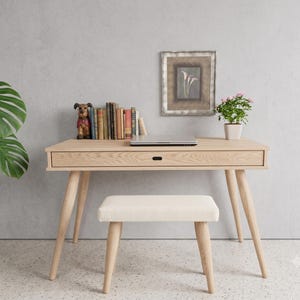 Könnte beinhalten: Ein heller Schreibtisch aus Holz mit einer einzelnen Schublade und vier konisch zulaufenden Beinen, dazu ein passender rechteckiger Hocker mit cremefarbener Polsterung. Auf dem Schreibtisch stehen ein geschlossener Laptop, Bücher und eine Hundefigur.