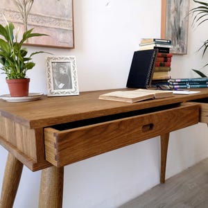Könnte beinhalten: Ein Schreibtisch aus Holz mit natürlicher Maserung, ausgestattet mit zwei Schubladen mit sichtbaren Schwalbenschwanzverbindungen und eingelassenen Griffmulden. Der Schreibtisch steht auf vier konischen Beinen und trägt eine grüne Pflanze, einen Bilderrahmen und Bücher.