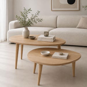 May include: A set of two light-toned wooden coffee tables with rounded edges and tapered legs. The taller oval table holds a ceramic vase with greenery, a small bowl, and two stacked books. The lower round table holds a small bowl and a book.