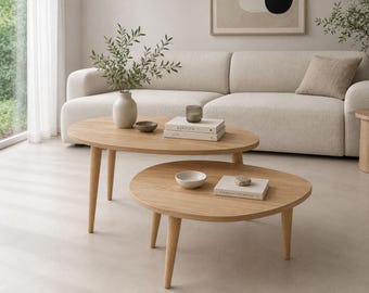 May include: A set of two light-toned wooden coffee tables with rounded edges and tapered legs. The taller oval table holds a ceramic vase with greenery, a small bowl, and two stacked books. The lower round table holds a small bowl and a book.