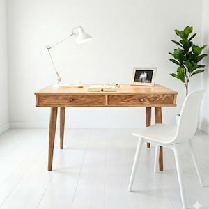 Peut inclure: Un bureau en bois clair avec deux tiroirs, une lampe de bureau blanche et une chaise blanche. Le bureau a une finition marron clair et est placé contre un mur blanc. Une photo encadrée, un livre et une tasse sont sur le bureau.