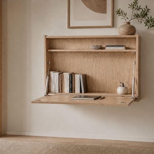 Op de afbeelding: Een aan de muur gemonteerd bureau van licht eikenhout met een neerklapbaar werkblad dat wordt ondersteund door twee hydraulische scharnieren van zilverkleurig metaal. De unit bevat een bovenplank met boeken en een kommetje, plus een laptop.