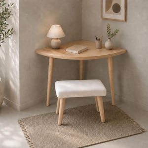 Puede incluir: Un escritorio esquinero de madera clara con borde frontal curvo y tres patas cónicas. Sobre él hay una pequeña lámpara blanca, un libro cerrado, un portalápices y un jarrón con vegetación. Incluye un taburete a juego con asiento blanco.